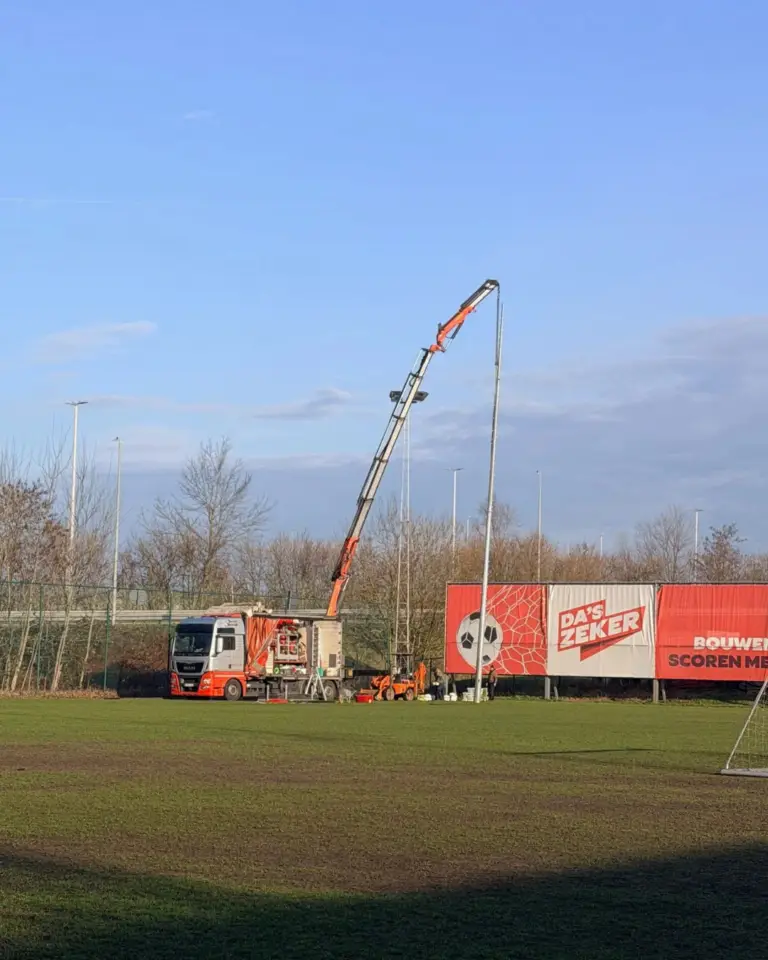 Hijskraan plaatst een nieuwe LED-verlichtingsmast op het voetbalterrein van K. Brecht SK tijdens de installatiefase.
