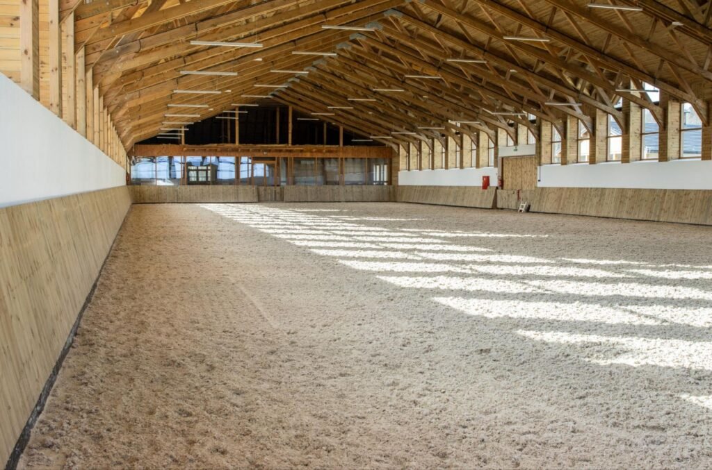 Lege overdekte paardenpiste met houten dakconstructie en zandbodem, verlicht met LED-verlichting.